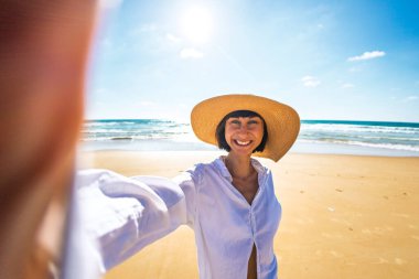 Hasır şapkalı, yaz tatilinin tadını çıkaran mutlu genç bir kadının yakın çekimi. Sahilde selfie çekip kameraya bakan güzel bir kızın portresi. Yaz yaşam tarzı.