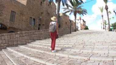 Sırt çantası ve kamera taşıyan bir kadın gezgin bir Arap şehrinin geniş basamaklarında yürür ve manzaranın fotoğrafını çeker. Seyahat