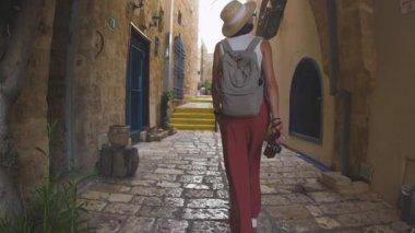 Kameralı bir kadın turist, şehrin eski turistik bölgesinde dar bir sokakta yürüyor ve Jaffa 'daki tatilinin tadını çıkarıyor.