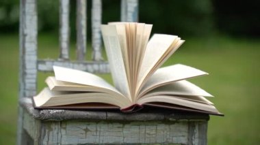Yazın yeşil bir çayırda eski küçük bir sandalyede rüzgarda sayfaları çeviren açık bir kitap. Yavaş çekim