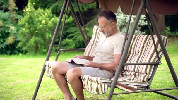 15歳の男は 休日に屋外の緑の晴れた夏の庭でスイングガーデンでスイングする本を読んでリラックスしています スローモーション — ストック動画