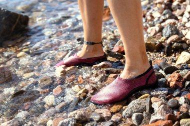 Su ayakkabısı giyen genç bir kadının ayakları taşlı bir sahilde dikiliyor. Keskin taşlara karşı koruma