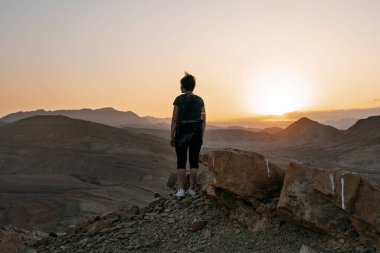 Sırt çantası takan kadın, günbatımında yüksek kayalıklarda tek başına duruyor ve İsrail 'deki Negev çölünün çöl dağlarında güneşin batışını seyrediyor.