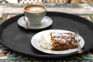 Kapuçino ve elmalı turtanın yakın çekimi. Süslü fayanslarla servis edilen bir açık hava restoranının tepsisinde çırpılmış kremayla servis ediliyor..