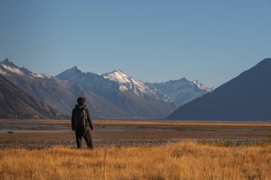 Genç adam güzel vadide duruyor. Arkasında dağlar ve sarı çayırlar var.