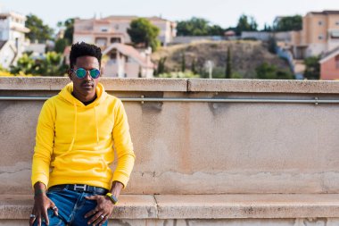 Yatay düzlem portresi. Sarı kazak giyen, mavi kot pantolon giyen ve gözlüklü genç bir siyah adam. Sokakta alçak bir duvara yaslanmış. Arka planda odak dışı evler görülebilir.