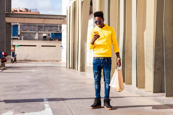 stock image A young black man dressed in a yellow sweatshirt, blue jeans and sunglasses using his smartphone while holding shopping bags. He is outside a shopping mall.