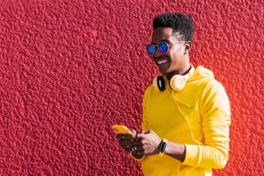 Sarı kazak ve güneş gözlüğü takmış gülümseyen genç Afrikalı bir adamın portresi cep telefonu ekranına dokunuyor ve pembe bir duvarın önünde boynuna kulaklık takıyor..
