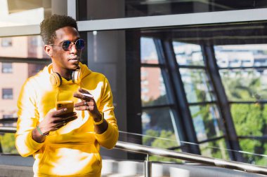 Sarı kazak ve güneş gözlüğü takmış, bir elinde sigara diğer elinde cep telefonu olan genç bir Afrikalı 'nın portresi. Boynunda bir kulaklık var..