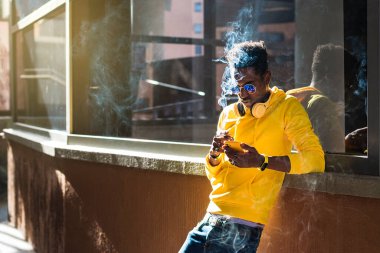 Sarı kazak, kot pantolon ve güneş gözlüğü giymiş genç Afrikalı bir adam bir pencere kenarına yaslanmış elinde sigara tutarken cep telefonuna bakıyor..