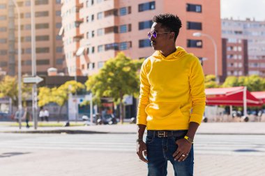 Sarı eşofman altı, kot pantolon ve güneş gözlüğü giymiş genç Afrikalı bir adamın portresi. Arka planda, sokakta duran binaları görebilirsiniz..
