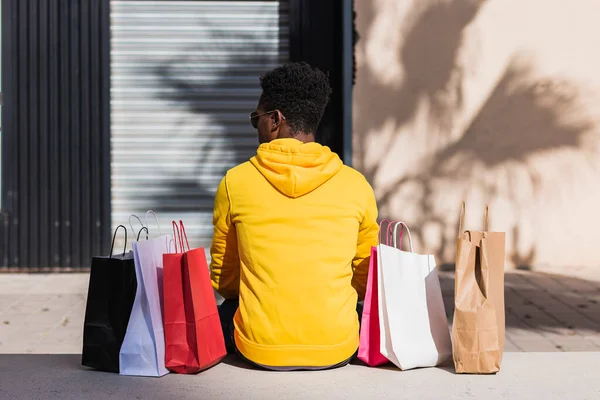 stock image A young African man dressed in a yellow sweatshirt sitting on a street bench next to some shopping bags seen from his back.