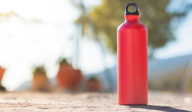 A red aluminum water bottle with black screw-on cap standing on a gray cement surface outside. The background is out of focus.