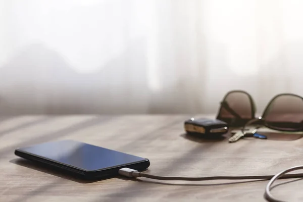 Mobile phone charging over a table with usb with cable. On background, defocused, there are car and home keys and sunglasses.
