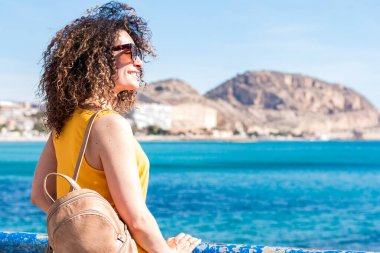 Sarı bluzlu neşeli, kıvırcık saçlı, güneş gözlüklü, rıhtımda duran ve gülümseyen genç bir kadın. Mavi gökyüzüne karşı dağlı, resimli deniz manzarasının tadını çıkarıyor.