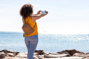 Sırt çantalı, kayalık sahilde duran ve yaz tatilinde akıllı telefondan dalgalanan denizin fotoğrafını çeken tanınmamış kıvırcık saçlı kadın turist.