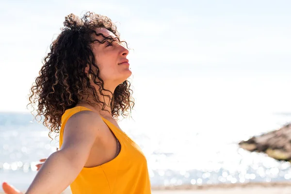 Side view of delighted woman with closed eyes spreading arms while standing on seashore in sunshine