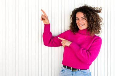 Kıvırcık kahverengi saçlı, parlak kazaklı, beyaz duvarın yanında duran ve kameraya bakarken kenara çekilen gülümseyen yetişkin bir kadın.