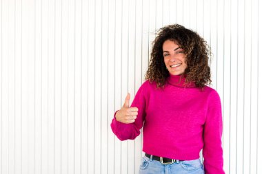 Pembe kazaklı, uzun kıvırcık saçlı, beyaz duvarın yanında duran ve kameraya bakarken jest yapan neşeli yetişkin kadın.