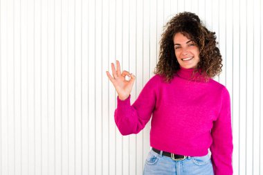 Pembe kazaklı, kıvırcık kahverengi saçlı, beyaz duvarın yanında duran ve kameraya bakarken işaret veren neşeli yetişkin kadın.