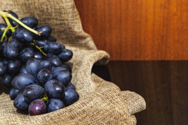 Yansıtıcı siyah bir yüzey ve ahşap bir arka plan üzerinde çuval bezle bir demet koyu üzümün yakın yatay fotoğrafçılığı.