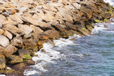 Denizin küçük dalgaları yığılmış bir grup taşa çarpıyor. Çarpıştıklarında su köpük üretir..