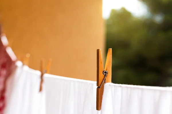 Blurred laundry colored clothes along with white hanging on clothesline while drying in sunlight and fastened with wooden clothespins against brown wall green trees and blue sky