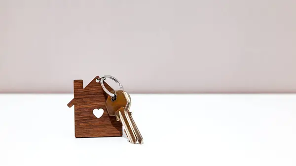 Home metallic keys in a wooden house-shaped keyring with a heart shape hole over a withe table in front of a light gray wall.