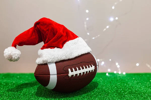 Amerikan futbol topu, gri bir duvarın yanında yeşil çimlerin üzerinde Noel Baba şapkası takıyor..