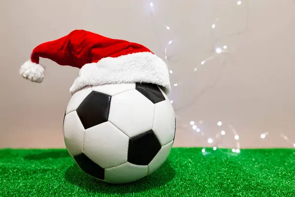 A black and white soccer ball wearing a Santa Claus red hat over green grass in front a light gray wall.