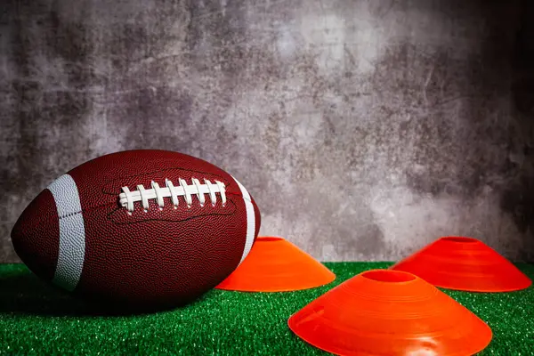 An american football ball and three orange cones over green grass ground in front of a gray concrete wall.