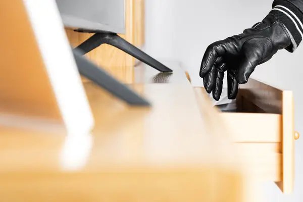 The hand of an unknown person wearing a black glove reaching into the drawer of a piece of furniture to steal its contents.