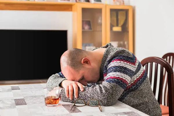 Kel ve yetişkin bir erkek, üzerinde alkollü içecek, buz ve bardaklar olan bir masanın üzerinde oturup yatıyor. Kişi üzgün, baş ağrısı ya da sarhoş görünüyor..