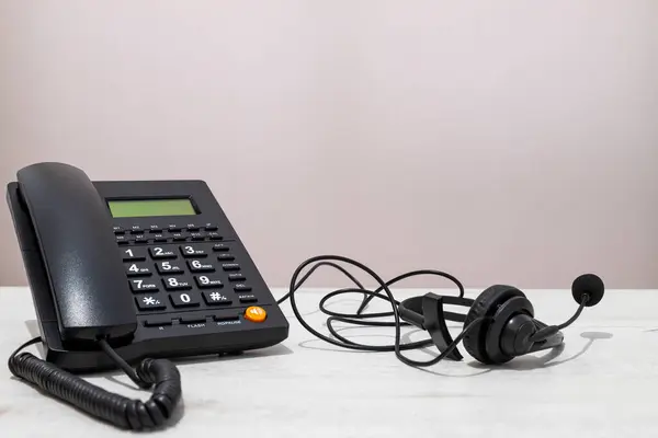 Kablosu sarkan siyah bir telefon. Kordonun üzerinde bir kulaklık var. Telefon masanın üstünde.