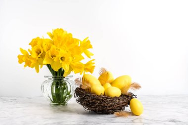 Yellow easter eggs in nest and bunch of daffodil flowers in vase on marble table and white background. Happy Easter and holiday preparation. Festive decoration.