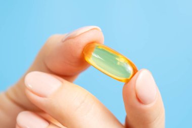 Omega 3 balık yağı kapsülünü tutan kadın elinin yakın çekimi. Mavi arka planda vitamin hapı ya da takviye. Sağlık ve bilimsel araştırmalar. Yaklaş, seçici odaklanma.