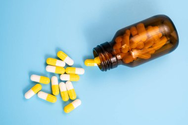 Beyaz ve sarı haplar mavi arka planda açılmış şişenin yanına saçılmış. İlaç, vitamin veya kapsüllerdeki takviye maddelerle tıbbi tedavi kavramı, uyuşturucu bağımlılığı.