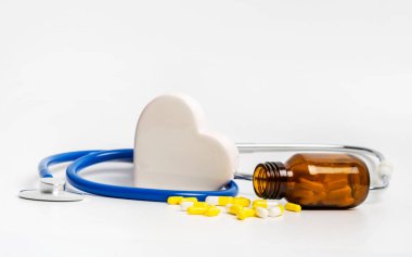 Cam şişenin yanında kalp hapları, seramik kalp ve beyaz arka planda steteskop. Kalp ve damar hastalıklarının kardiyoloji tedavisi için ilaç, diyet takviyeleri ve kalp sağlığı için vitaminler.