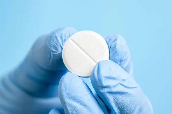 White effervescent or carbon tablet in hand in disposal glove. Doctor, pharmacist or scientist fingers holding pill, vitamin or supplement on blue background. Medicine therapy, scientific research.
