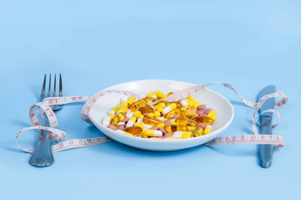 Many colorful pills, omega 3 vitamins and nutrition supplements in white plate, fork, knife and measuring tape on blue background. Diet and weight loss medicine. Medical treatment, weight loss drugs.