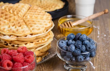 Bir yığın Belçika waffle 'ı, bal ve ahududu ve yabanmersinli kase. Lezzetli yaz kahvaltısı. Arka planda ev yapımı yuvarlak waffle ve waffle makinesi var..