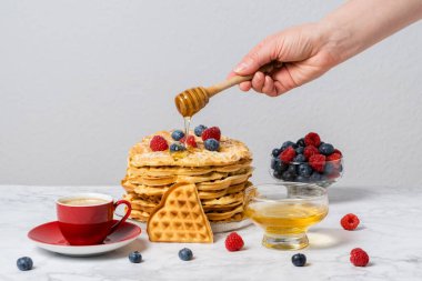 Kadın eli Belçika waffle 'larına bal döküyor. Kalp şeklinde waffle 'lar, taze böğürtlenler ve mermer masada bir fincan kırmızı kahve ile tatlı bir yaz kahvaltısı..