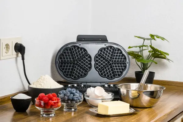 Stock image Ingredient for Belgian waffles and waffle maker on kitchen table. Bowl with dough, cracked eggs, butter, sugar, flour and berries. Making waffles at home.