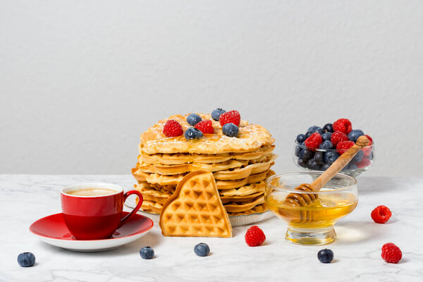Stack of Belgian waffles, berries, honey and red cup of coffee: delicious summer breakfast on marble table. Heart-shaped waffle and mix of raspberries and blueberries as topping.
