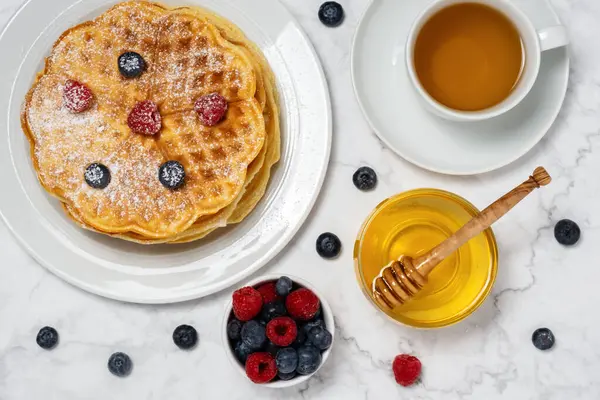 stock image Belgian waffles, berries, honey and cup of tea: delicious summer breakfast from above. Heart-shaped waffle and mix of raspberries and blueberries as topping on marble background. Flat lay, top view