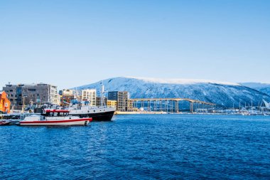 Kışın Tromso limanı, Norveç. Tromso limanındaki Sandnessundet Köprüsü ve arka planda dağlar olan teknelerin görüntüsü. Norveç 'in kuzeyindeki Kuzey Kutup Dairesi kasabasında karlı kış manzarası.