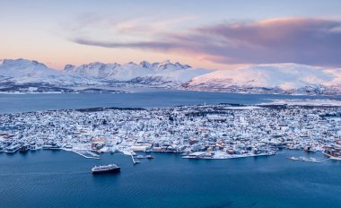 Tromso, Norveç 'in hava manzarası. Storsteinen Dağı' ndan kışın gün batımında. Tromsoya adası, şehir merkezindeki karlı çatıları, limanı, fiyort suyundaki yolcu gemisi ve arka plandaki dağlarıyla.