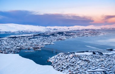 Tromso, Norveç 'in hava manzarası. Storsteinen Dağı' ndan kışın gün batımında. Tromsoya adası, Tromso şehir merkezi, liman, Fiyort üzerindeki Sandnessundet Köprüsü ve arka plandaki dağlarla karlı çatılar..