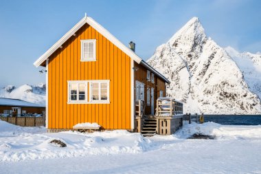 Geleneksel sarı rorbu evi ve Norveç 'in Lofoten adalarındaki Sakrisoy balıkçı köyünün Olstind dağı zirvesi. Karlı dağlarla ve İskandinav kulübeleriyle kış manzarası.