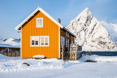 Traditional yellow rorbu house and Olstind mountain peak in Sakrisoy fishing village on Lofoten islands, Norway. Winter landscape with snowy mountains and Scandinavian cottage. clipart